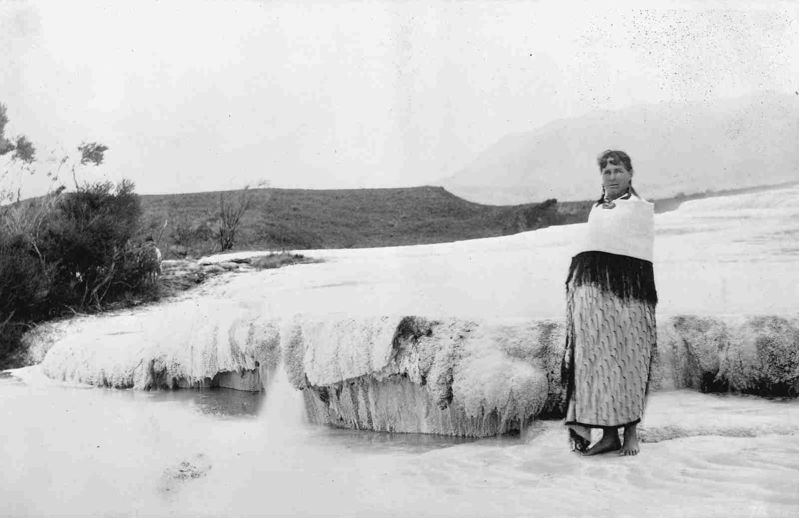 Black and white photograph of guide Sophia Hinerangi standing barefoot on a white silica terrace at Lake Rotomahana in 1885, wrapped in a shawl, hills behind