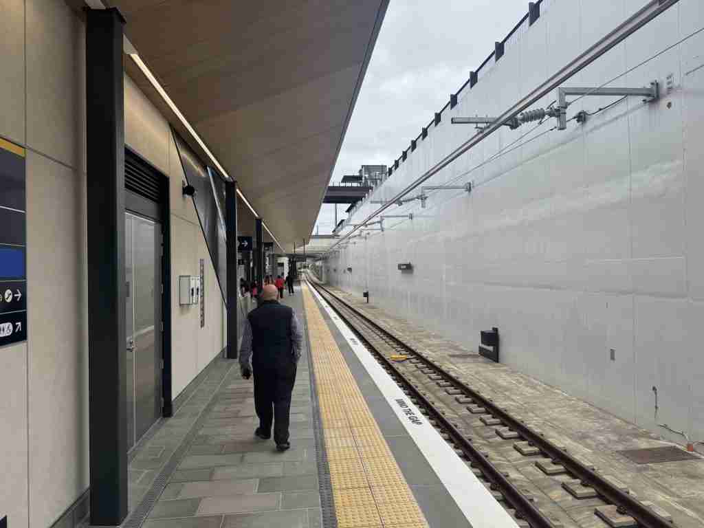 A passenger walking along the open-air platform at Maungawhau Station, with concrete retaining walls and overhead wires